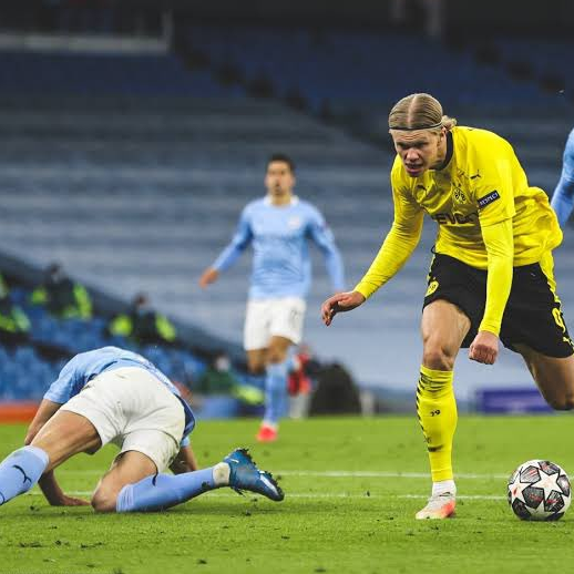 Piłkarz Borussii Dortmund prowadzi piłkę obok leżącego rywala piłkarza Manchesteru City podczas dynamicznej akcji meczu.